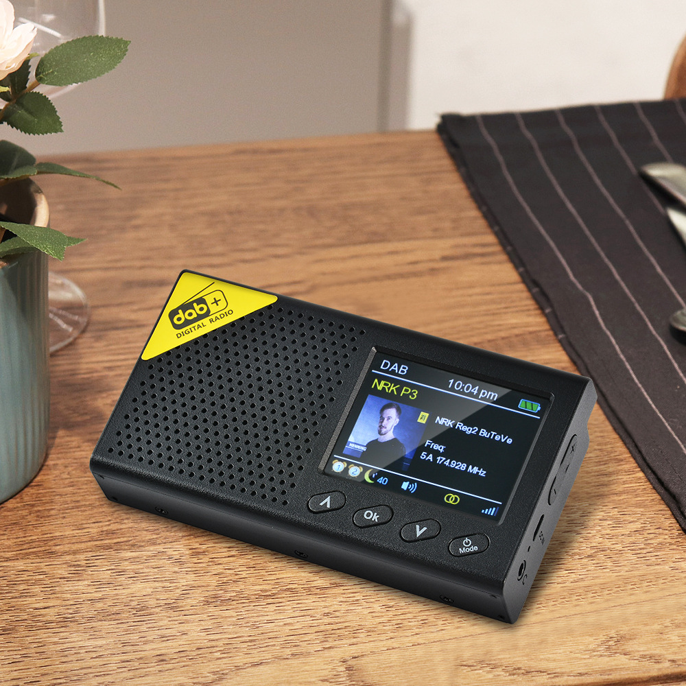 Ein schwarzes DAB+-Digitalradio liegt auf einem Holztisch. Der Bildschirm zeigt ein Männerbild und Radioinfos. Daneben liegt eine schwarze Serviette.