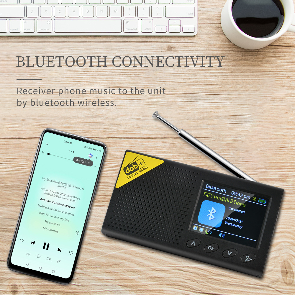 Ein Telefon spielt Musik, verbunden mit einem schwarzen Radio. Eine Tastatur und eine Tasse Kaffee stehen auf einer Holzoberfläche.