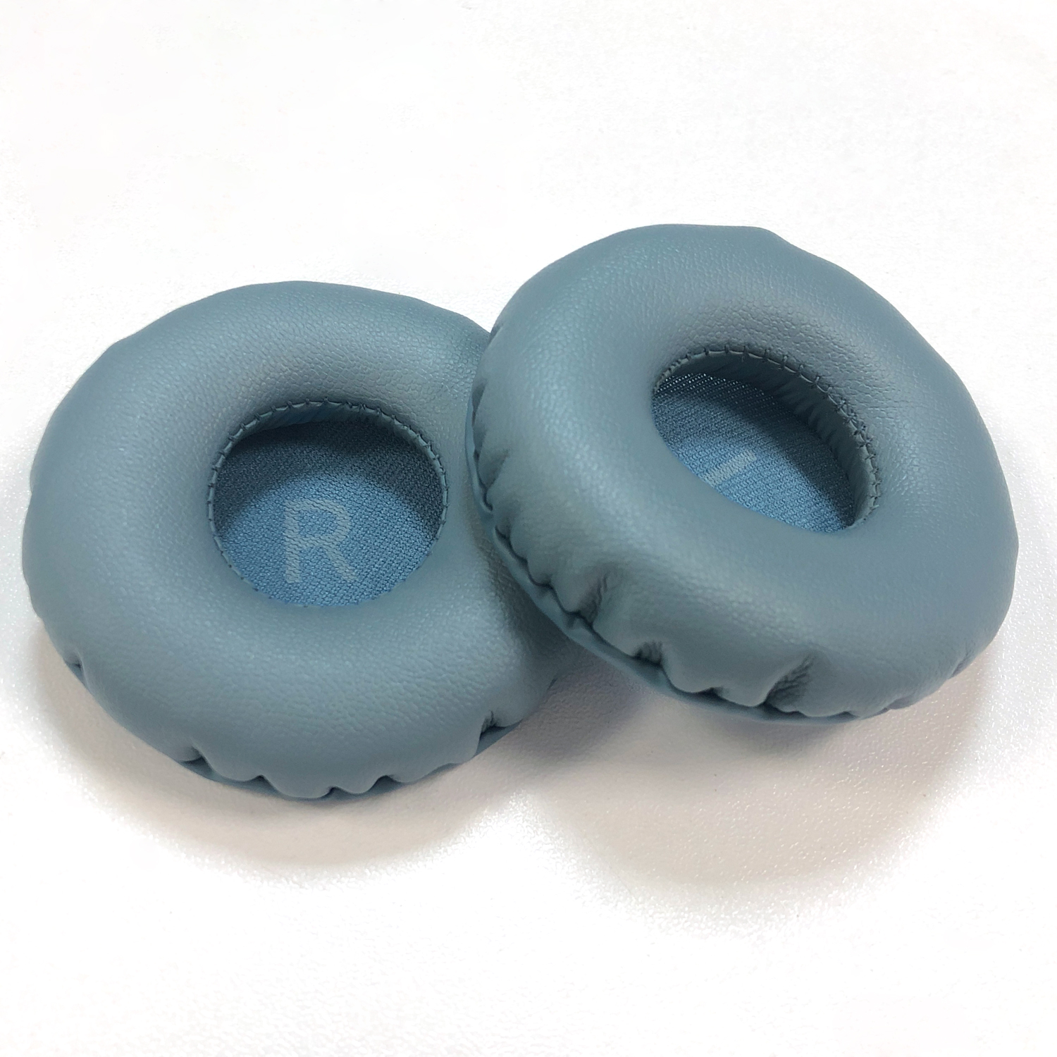 Dwie niebieskie nauszniki na białej powierzchni. Nauszniki są okrągłe z wewnętrznym wypełnieniem. Na jednym 'R'.