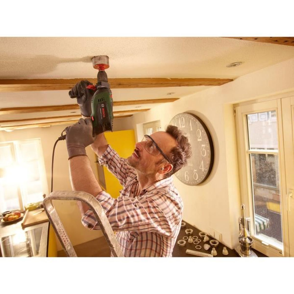 Mężczyzna wierci w suficie. Używa wiertarki stojąc na drabinie. W tle zegar i okno.