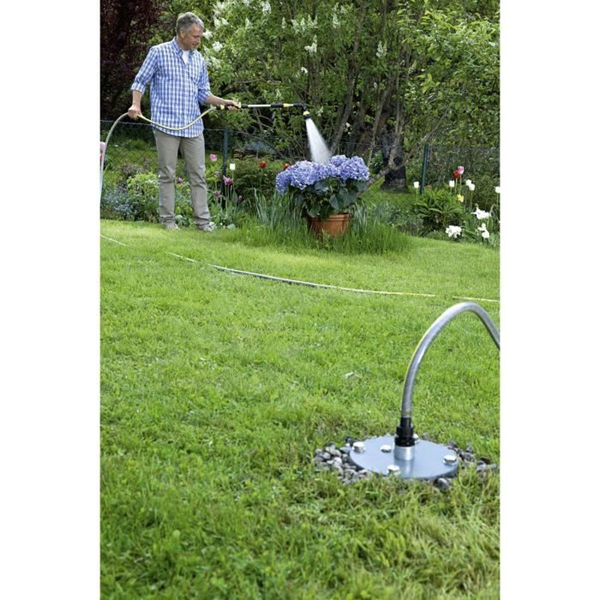 Hombre regando flores azules en maceta con manguera. Césped verde llena el jardín. Aspersor en primer plano.