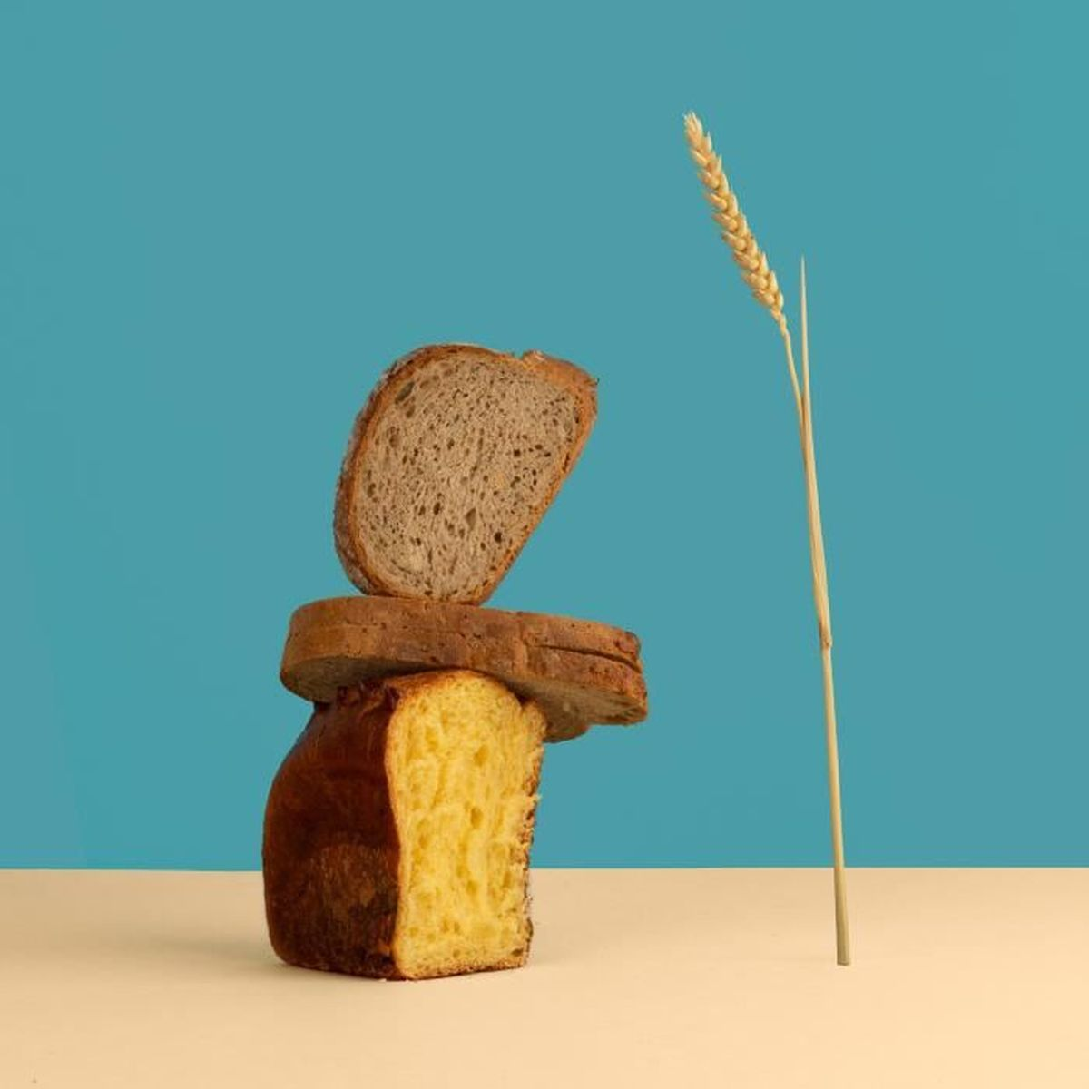 Ułożone na stosie kromki chleba, ze źdźbłem pszenicy. Górna kromka jest brązowa, środkowa ciemniejsza, dolna żółta. Tło niebieskie i kremowe.