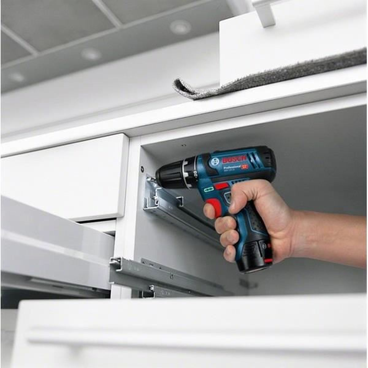 Eine Hand hält einen blau-schwarzen Bosch-Bohrer und schraubt in einen weißen Schrank. Die Szene ist in einer Küche mit weißen Schränken.
