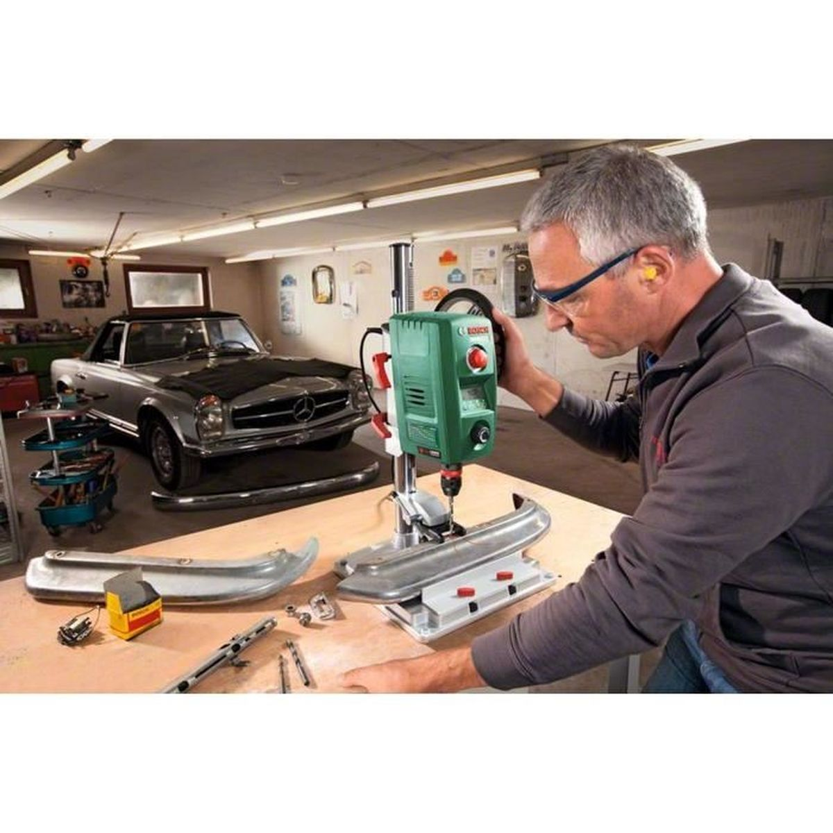 Ein Mann benutzt eine Ständerbohrmaschine in einer Garage. Im Hintergrund ist ein Oldtimer zu sehen. Autoteile und Werkzeuge liegen auf dem Tisch.