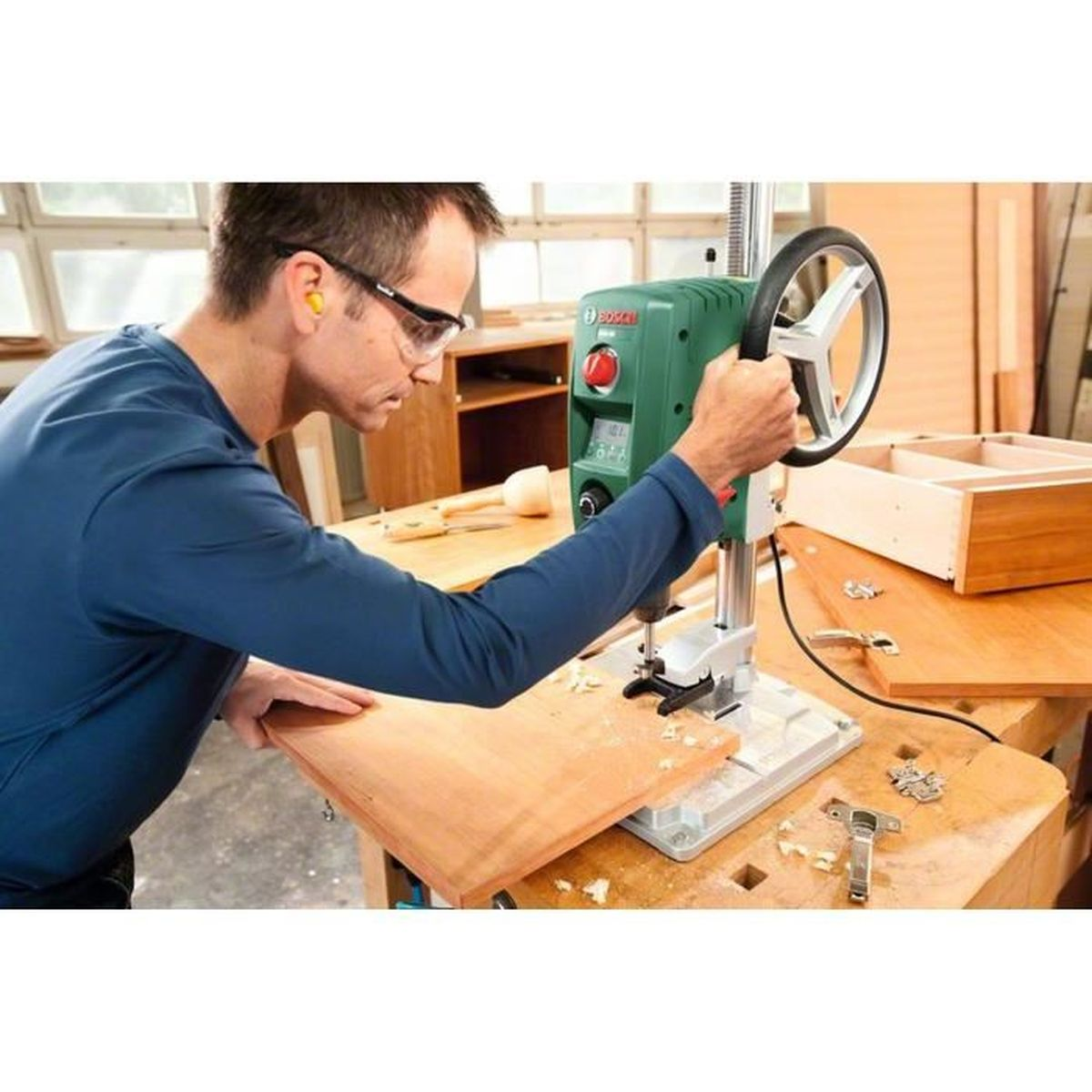 Ein Mann mit Schutzbrille benutzt eine Bohrmaschine an einer Holzplatte. Sägemehl, Werkzeuge und eine teilweise gebaute Schublade sind sichtbar.