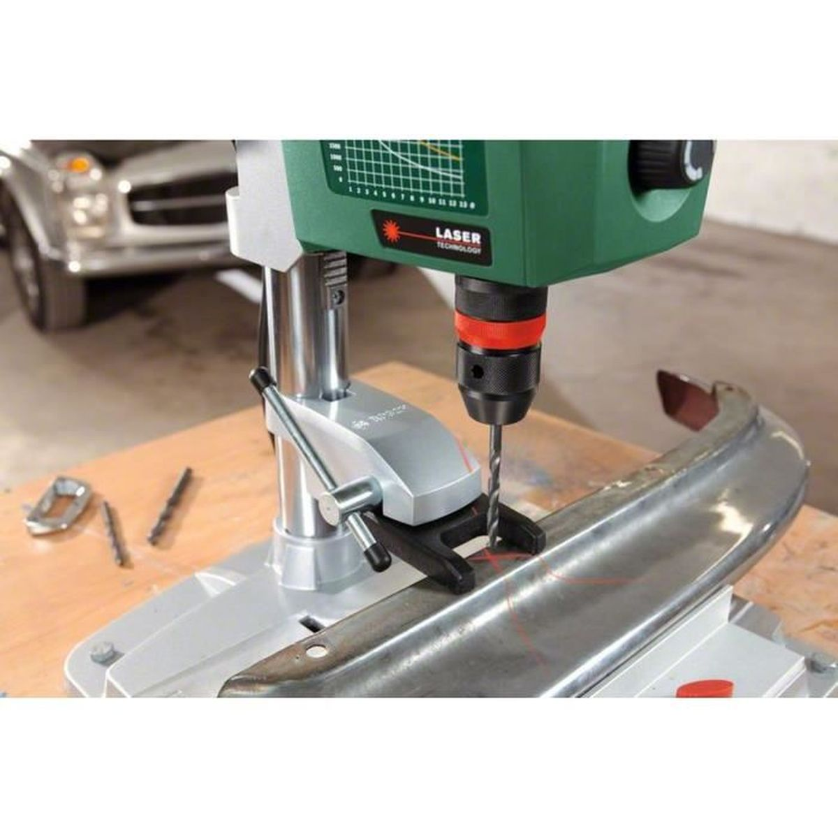 Eine Bohrmaschine bohrt ein Metallobjekt. Die Bohrmaschine ist grün und schwarz. Das Metallobjekt ist silberfarben. Im Hintergrund ist ein Auto zu sehen.