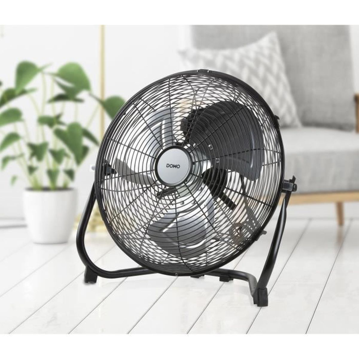 Un ventilateur DOMO noir est sur un sol blanc. Une plante en pot et une chaise grise sont en arrière-plan.