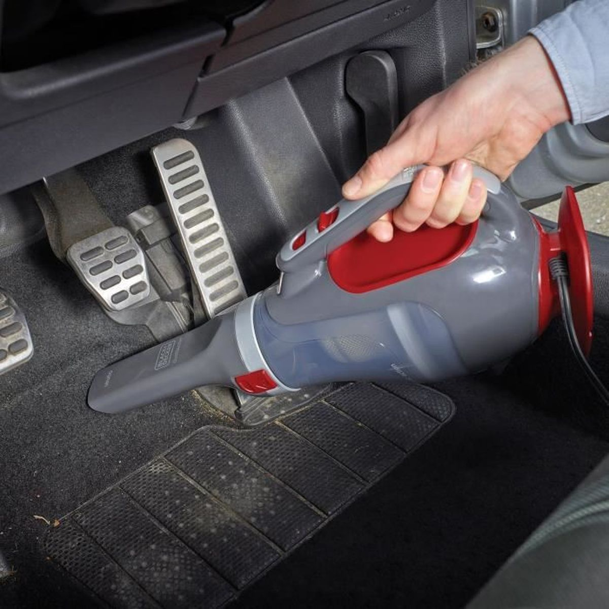 Ręka trzyma szaro-czerwony odkurzacz ręczny w samochodzie, czyszcząc czarną podłogę. Widoczne są pedały.
