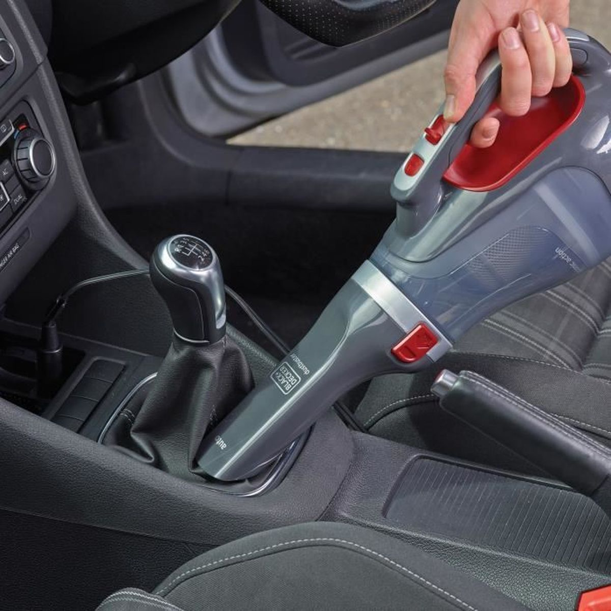 Osoba używa odkurzacza ręcznego do czyszczenia wnętrza samochodu. Odkurzacz jest szary i czerwony. Samochód ma dźwignię zmiany biegów i hamulec ręczny.