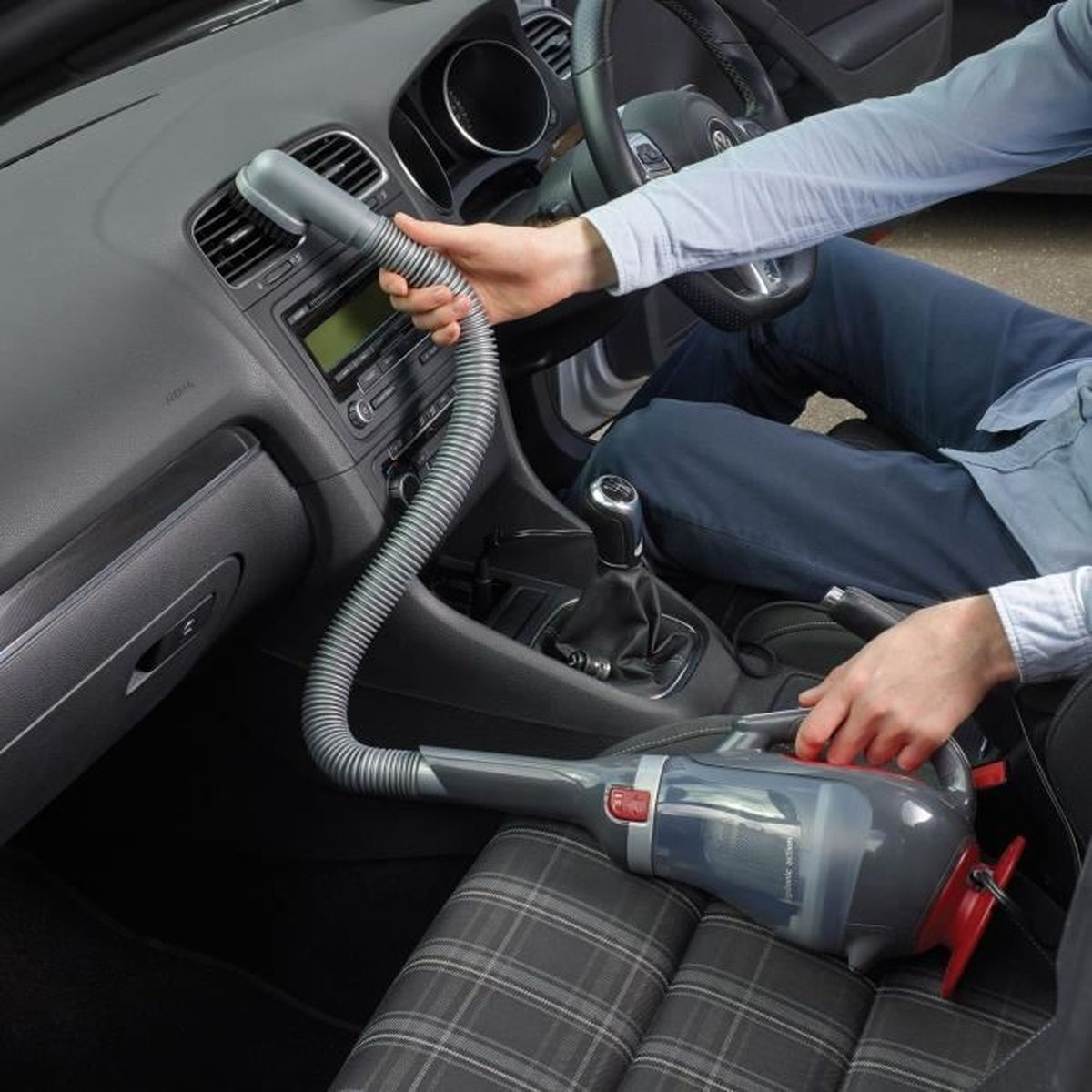 Osoba odkurza wnętrze samochodu. Odkurzacz ręczny jest używany na desce rozdzielczej i siedzeniach. Widoczna jest kierownica i dźwignia zmiany biegów.