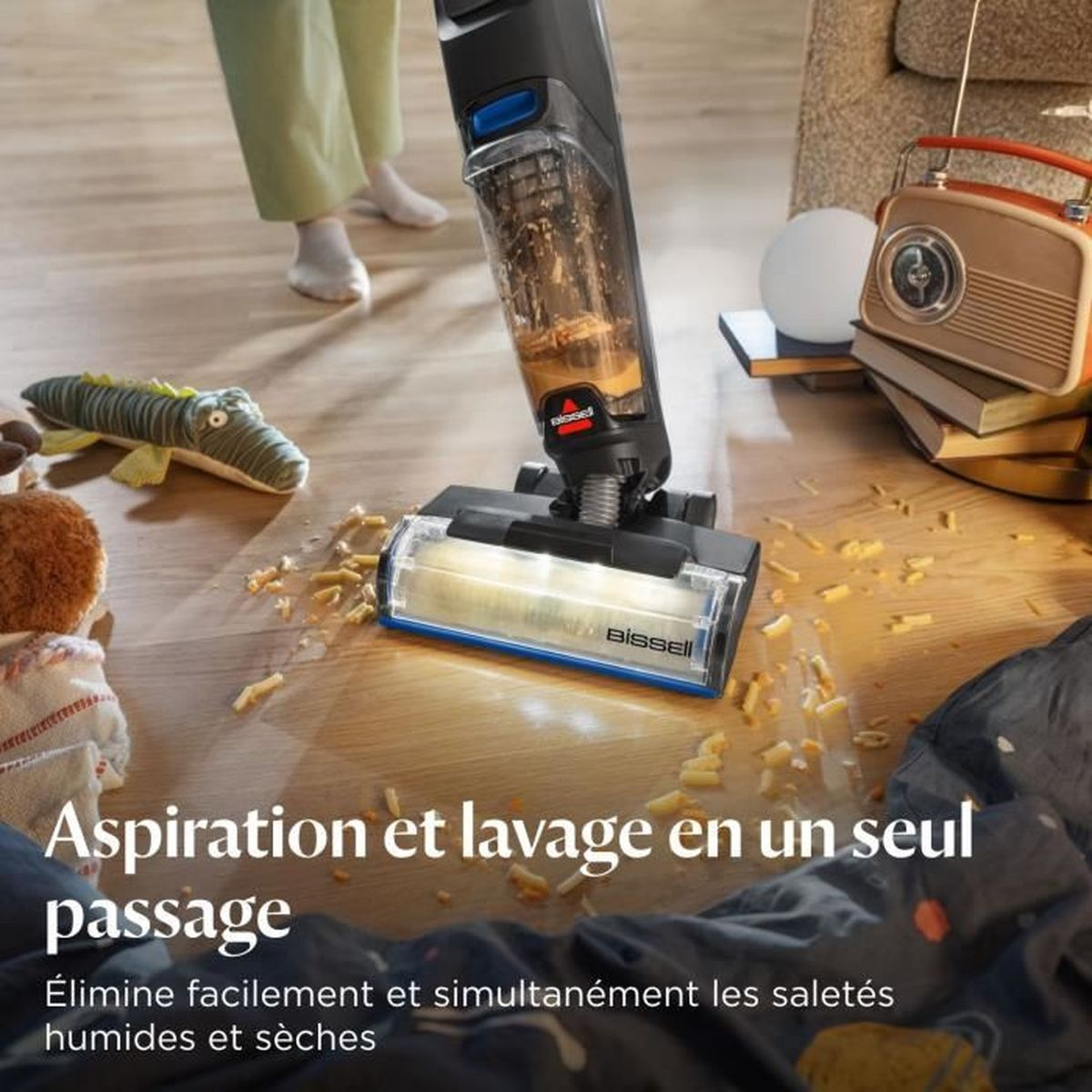 Ein Staubsauger reinigt einen Holzboden. Spielzeug, ein Radio und Bücher sind im Hintergrund. Der Text lautet 'Aspiration et lavage en un seul passage'.