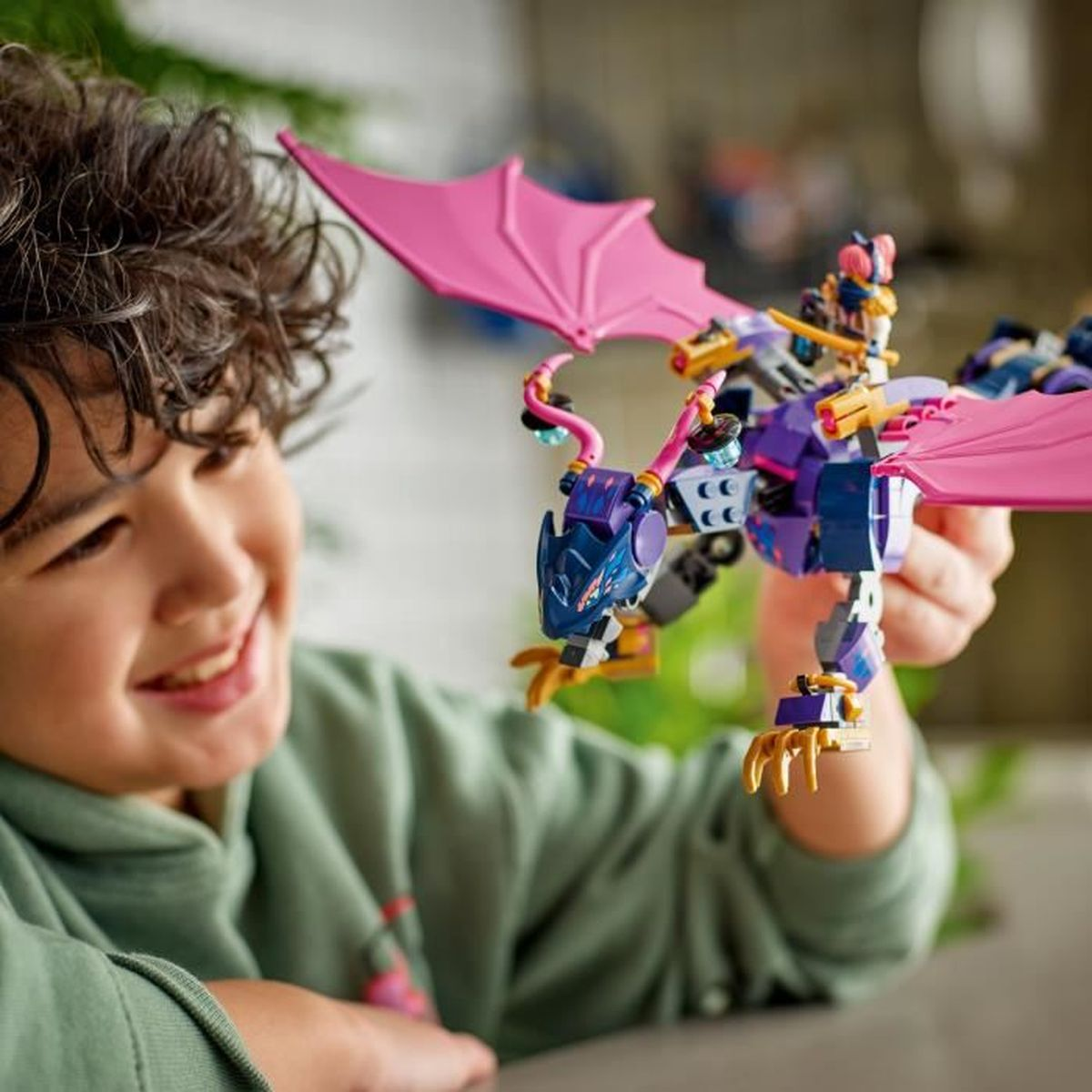 Dziecko trzyma smoka Lego z różowymi skrzydłami. Smok jest fioletowy i niebieski. Dziecko się uśmiecha. Tło jest rozmyte.