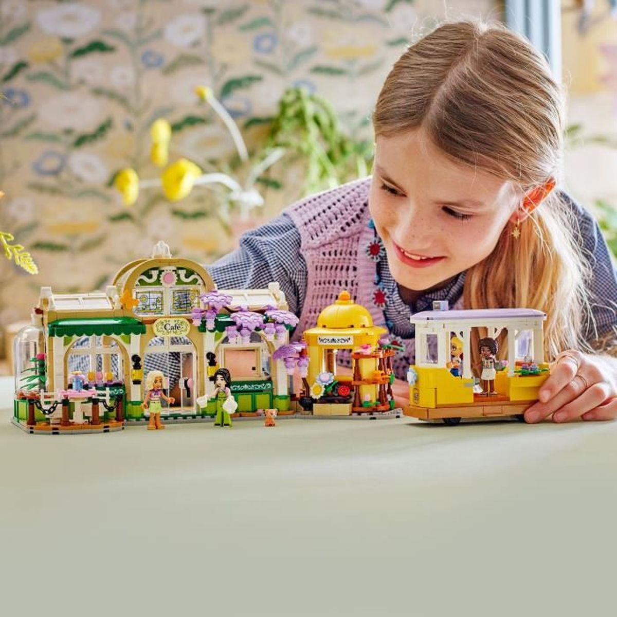 Dziewczyna bawi się zestawem Lego, w którym znajduje się kawiarnia, sklepy i żółty tramwaj. Zestaw stoi na zielonej powierzchni.