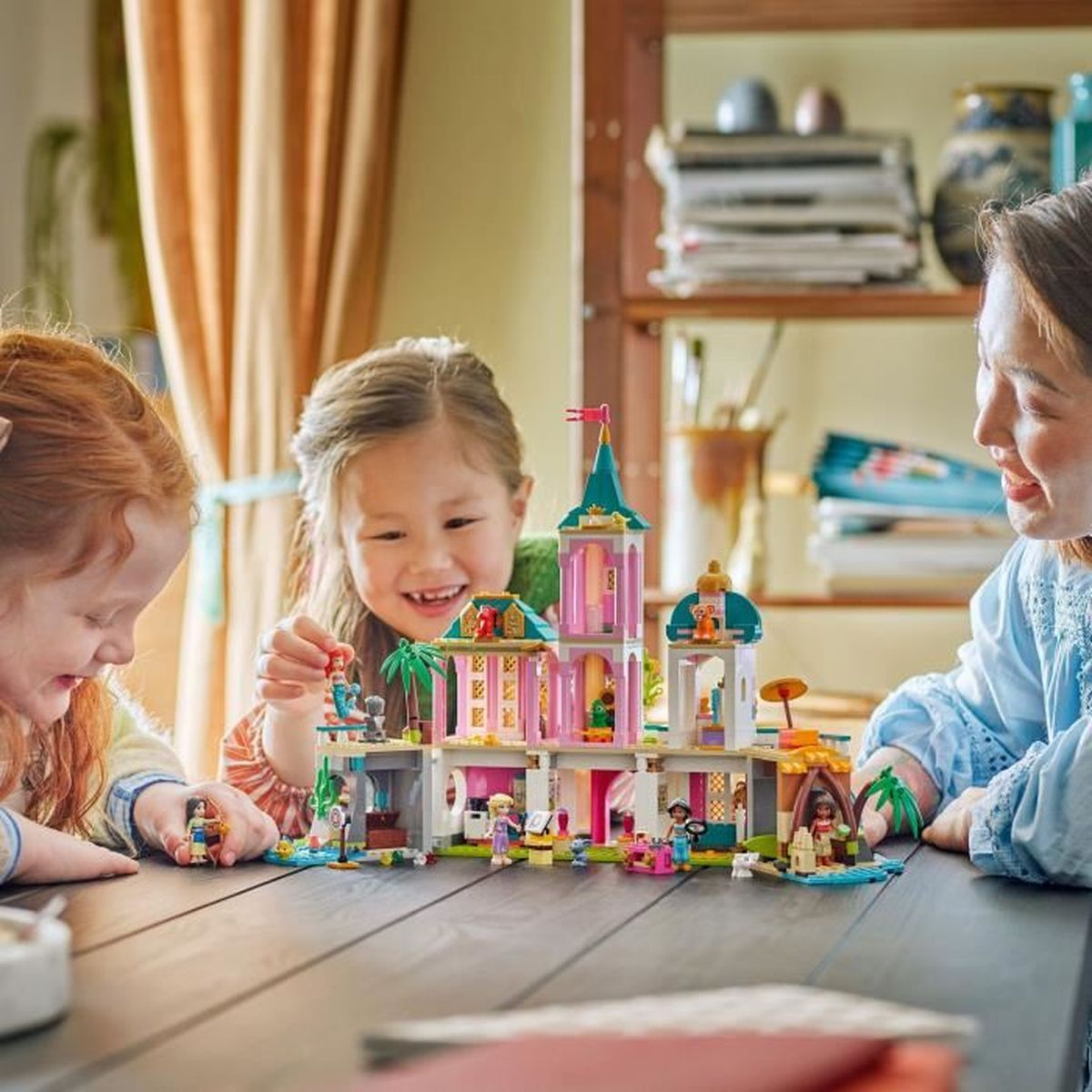 Tres personas juegan con un castillo de Lego sobre una mesa. El castillo es rosa y blanco. La gente sonríe.