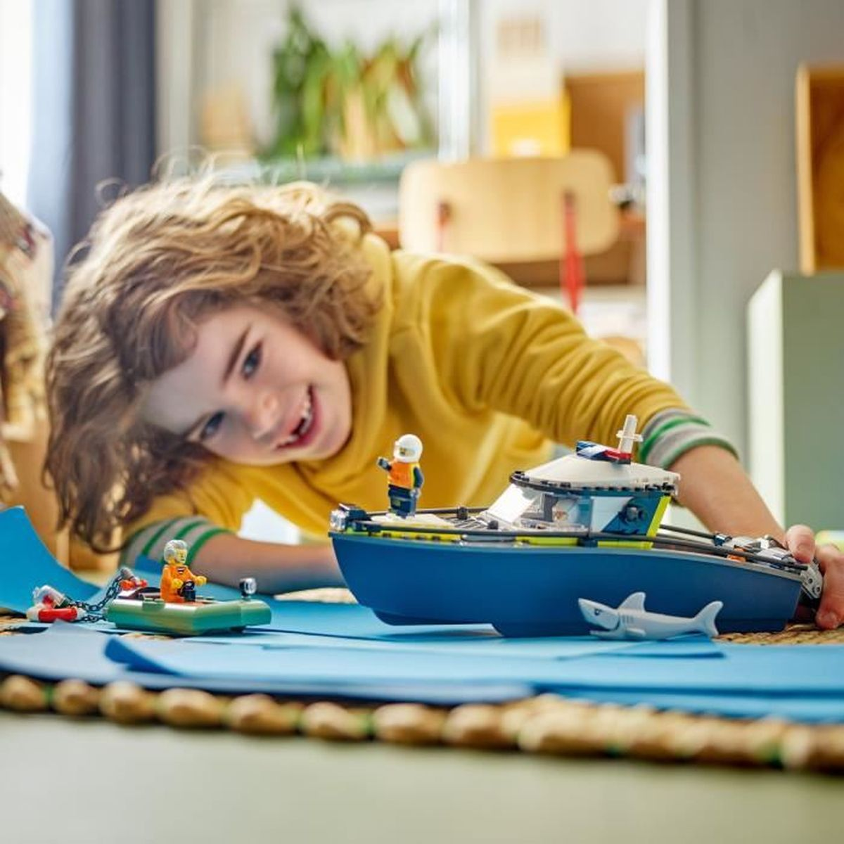 Dziecko w żółtej bluzie z kapturem uśmiecha się, bawiąc się zabawką łodzią i małą łódką. Zabawkowa rekin jest w pobliżu łodzi.
