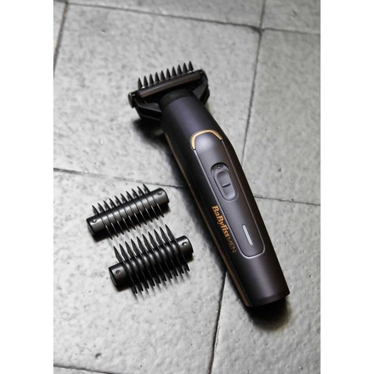 Ein schwarzer und goldener Babyliss Herren-Trimmer mit zwei Aufsätzen auf einer grauen Fliesenoberfläche.