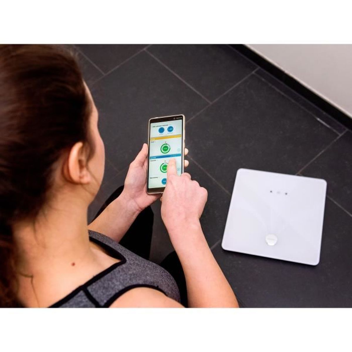 Una donna usa uno smartphone per controllare i dati, una bilancia è sul pavimento. La donna indossa un top grigio. Piastrelle nere.