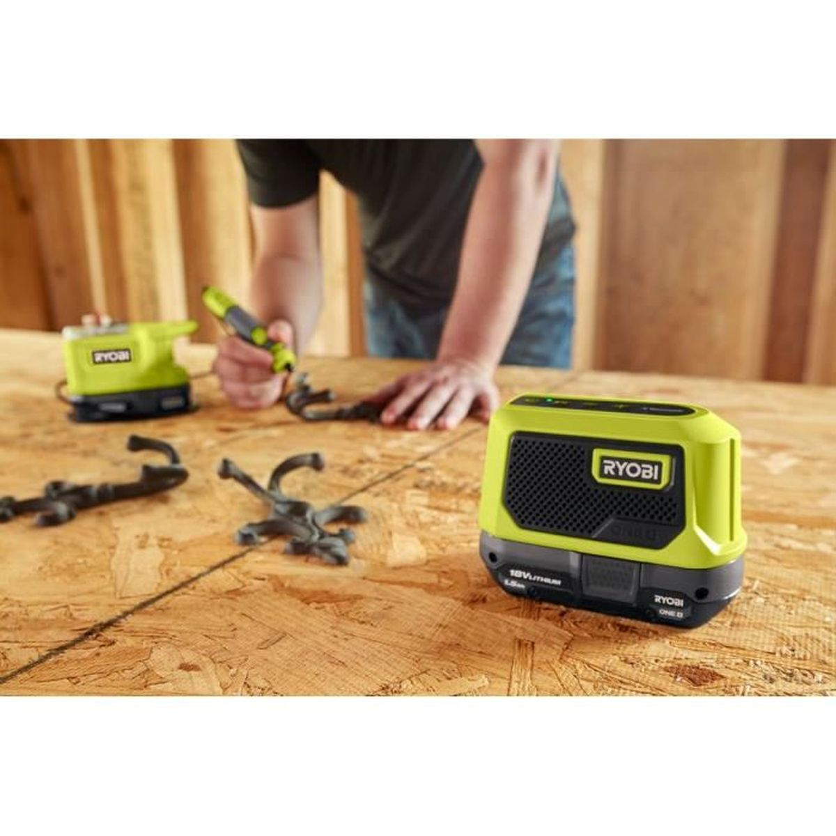 Una persona usa uno strumento su una superficie di legno. Sono presenti diversi strumenti Ryobi e oggetti di metallo scuro. Lo sfondo mostra pannelli di legno.