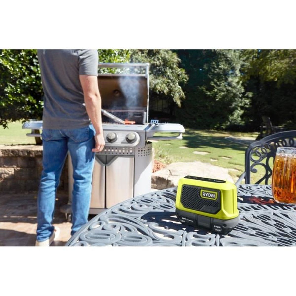 Un uomo fa un barbecue all'aperto, vicino a un tavolo con un altoparlante Ryobi e una bevanda. Alberi verdi sullo sfondo.