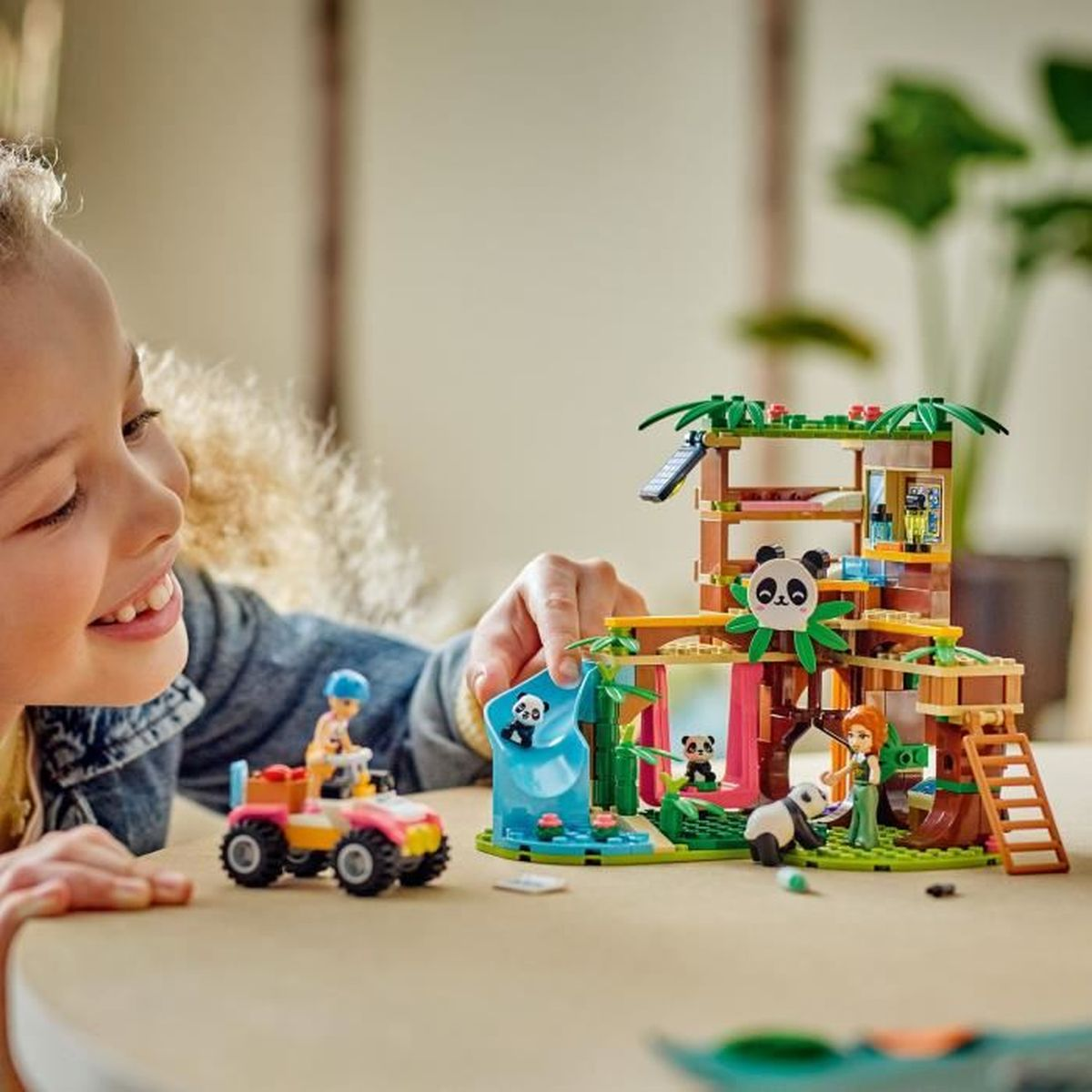 Dziecko uśmiecha się, bawiąc się zestawem Lego z pandami. Zestaw zawiera domek na drzewie, pandy i mały pojazd.