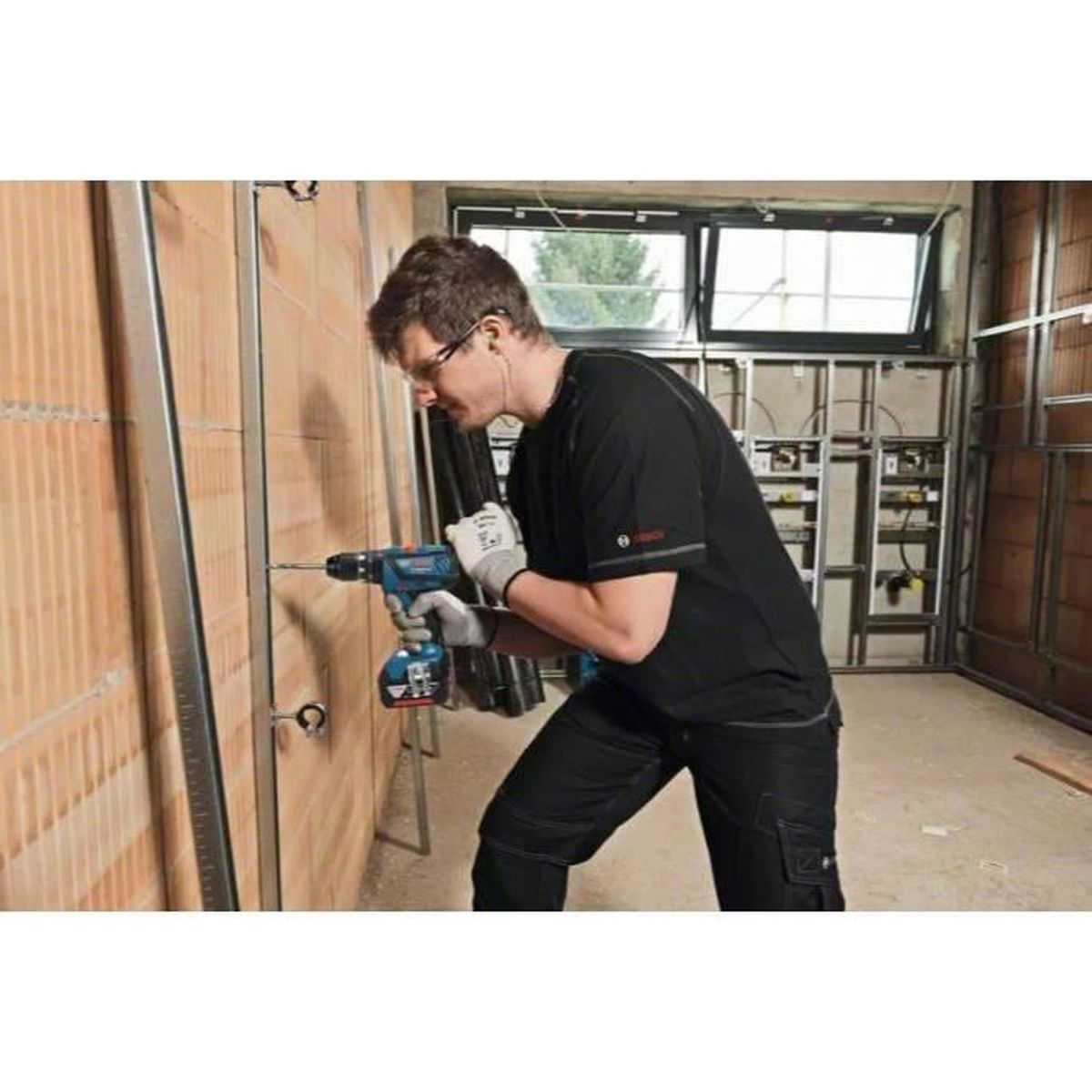 Ein Mann in schwarzer Arbeitskleidung bohrt in eine Wand. Er trägt Schutzbrille und Handschuhe. Die Wand ist aus Ziegeln.