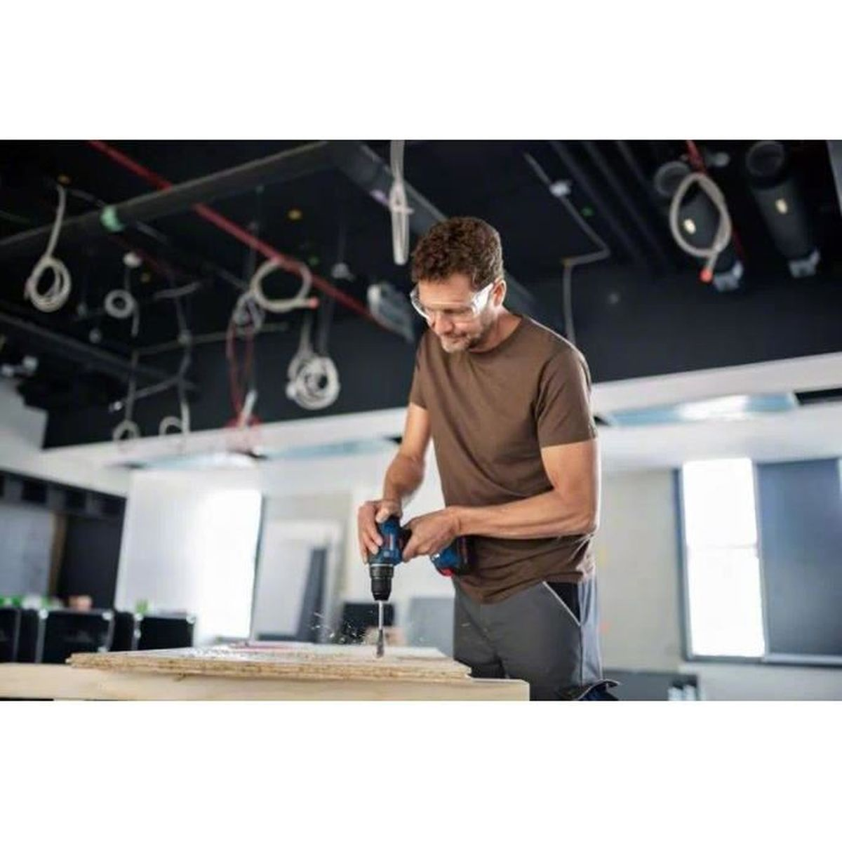 Mężczyzna w okularach ochronnych wierci w drewnie. Ma na sobie brązową koszulę i szare spodnie. Nad nim są przewody i światła.