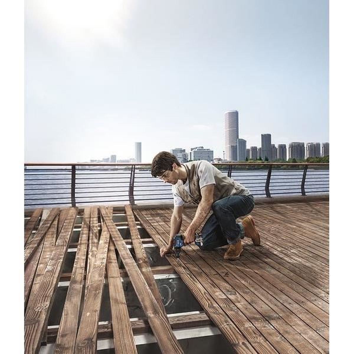 Mężczyzna klęczy, używając elektronarzędzia na drewnianych deskach. Panorama miasta i woda w tle. Dominują brązowe i niebieskie tony.