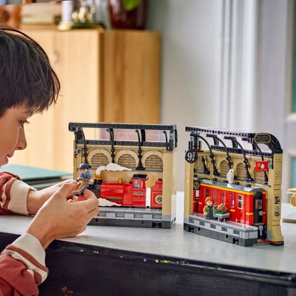 Un niño juega con sets de estación de tren LEGO, con trenes rojos, personajes y el letrero 9 3/4. Un armario de madera está en el fondo.