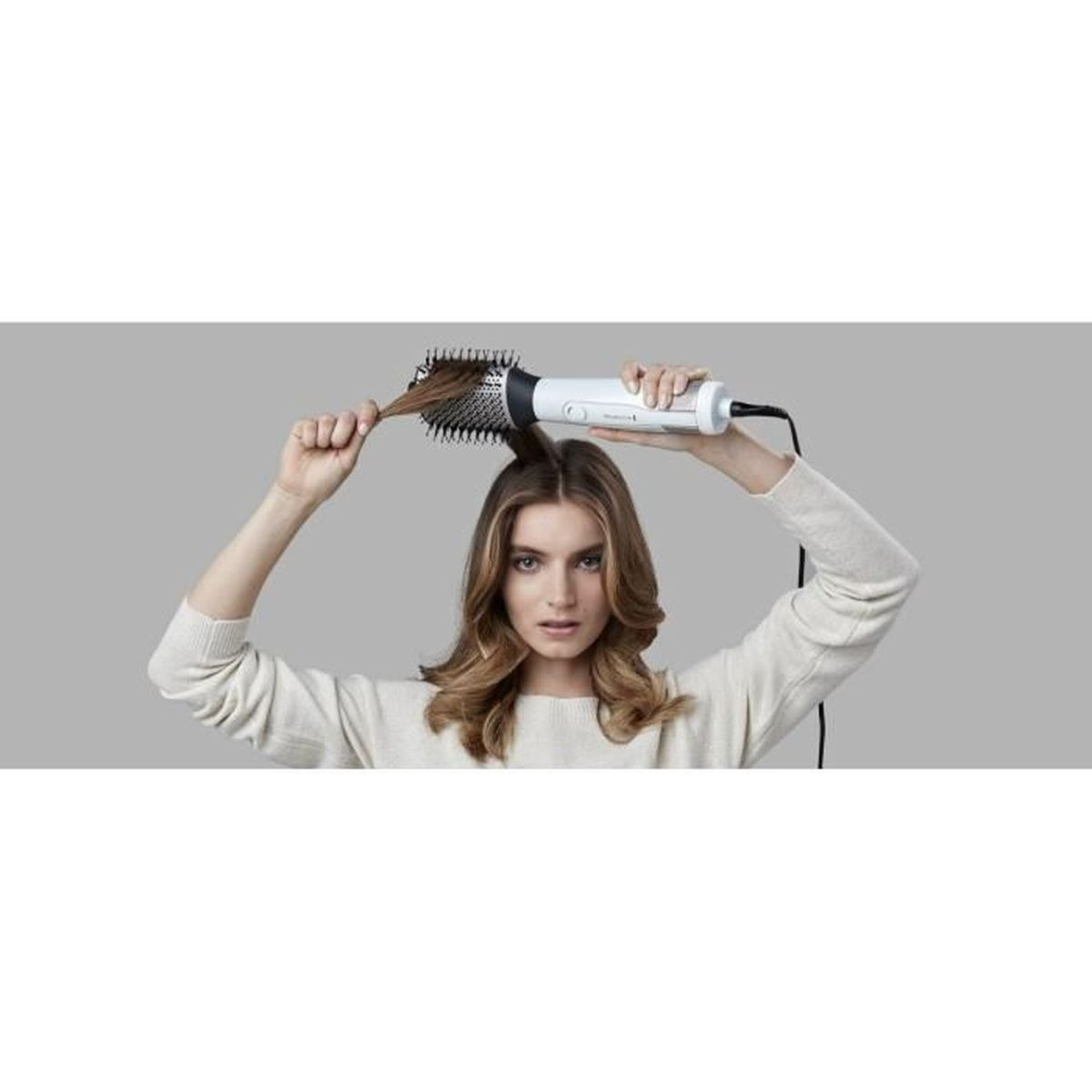 Kobieta stylizuje włosy biało-czarną szczotką do włosów. Trzyma ją nad głową. Tło jest szare.