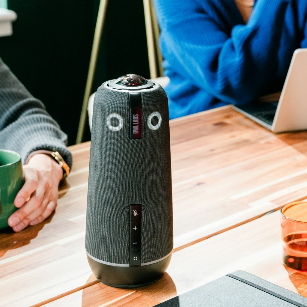 Ein dunkelgraues Owl Labs-Gerät für Besprechungen auf einem Holztisch. Menschen sitzen am Tisch. Ein Laptop und ein Notizbuch sind zu sehen.