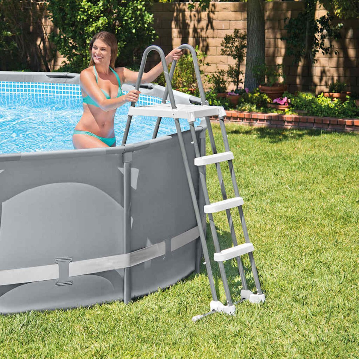 Frau im türkisfarbenen Bikini im Pool, hält eine Leiter. Pool ist grau, umgeben von Gras. Ziegelmauer im Hintergrund.