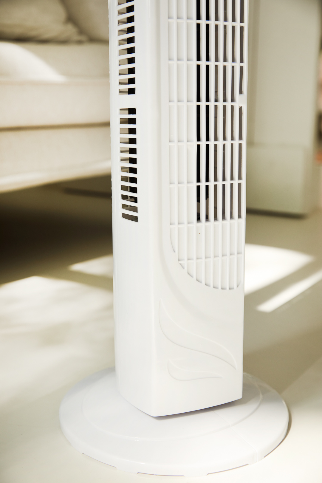 A white tower fan stands on a white floor. The fan is rectangular with vertical slats. Background shows a light-colored surface.