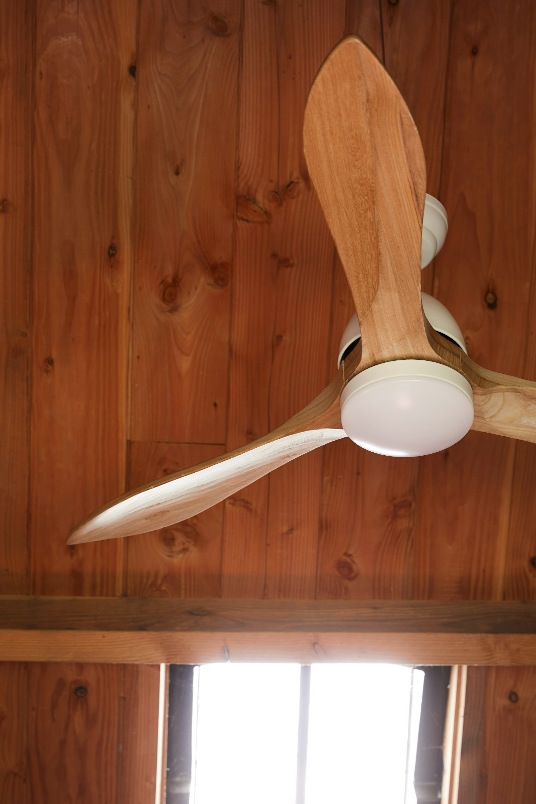 Ventilatore da soffitto in legno con tre pale. Pale in legno chiaro e alloggiamento motore bianco. Soffitto in legno e finestra luminosa.