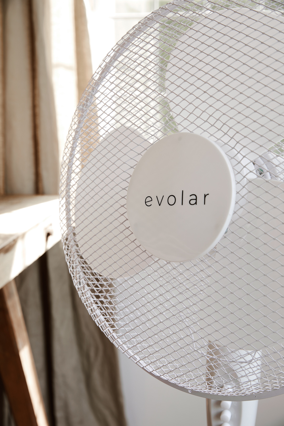 A white electric fan with a protective mesh. The brand name 'evolar' is visible. Background includes curtains and a table.
