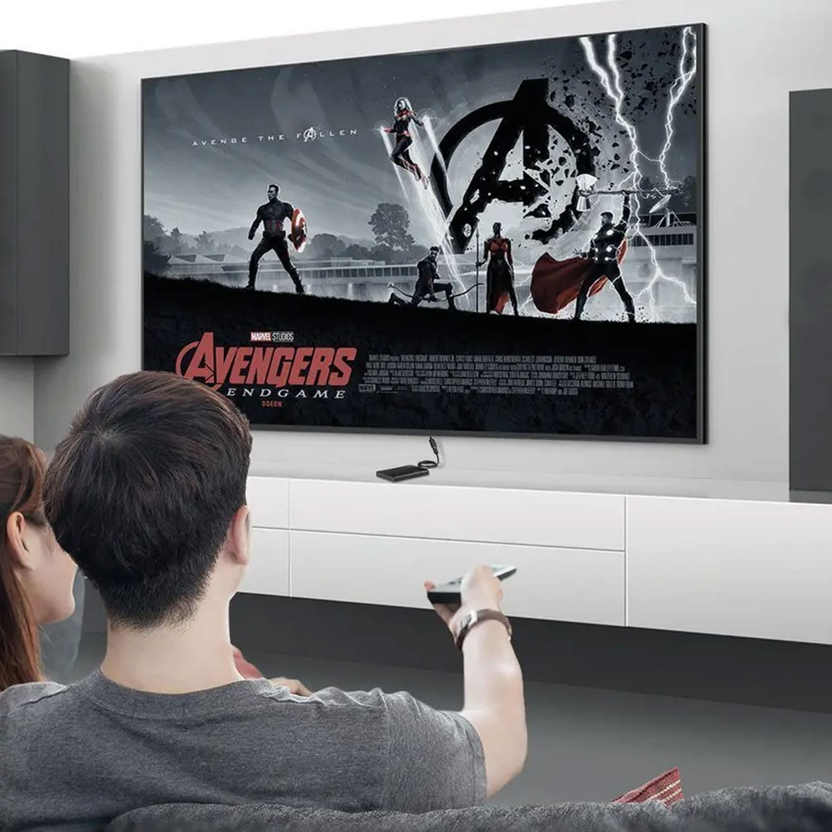 Para ogląda film Avengers Endgame na dużym ekranie telewizora. Plakat filmowy jest widoczny. Osoba trzyma pilota.