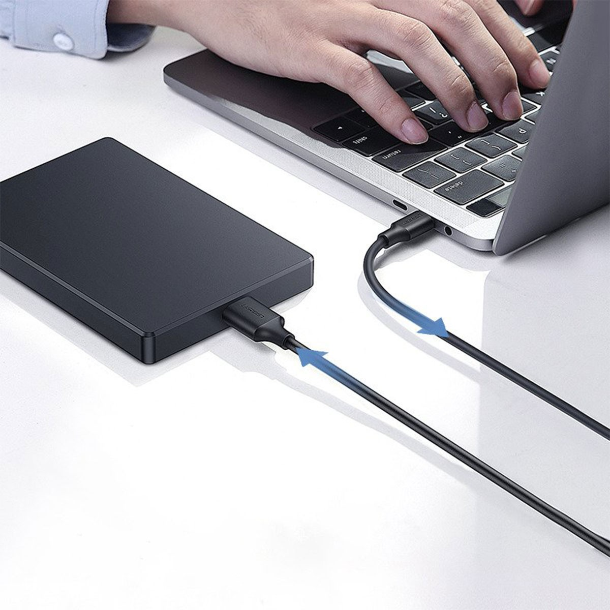 Czarny zewnętrzny dysk twardy jest podłączony do laptopa czarnym kablem. Ręka pisze na klawiaturze laptopa.