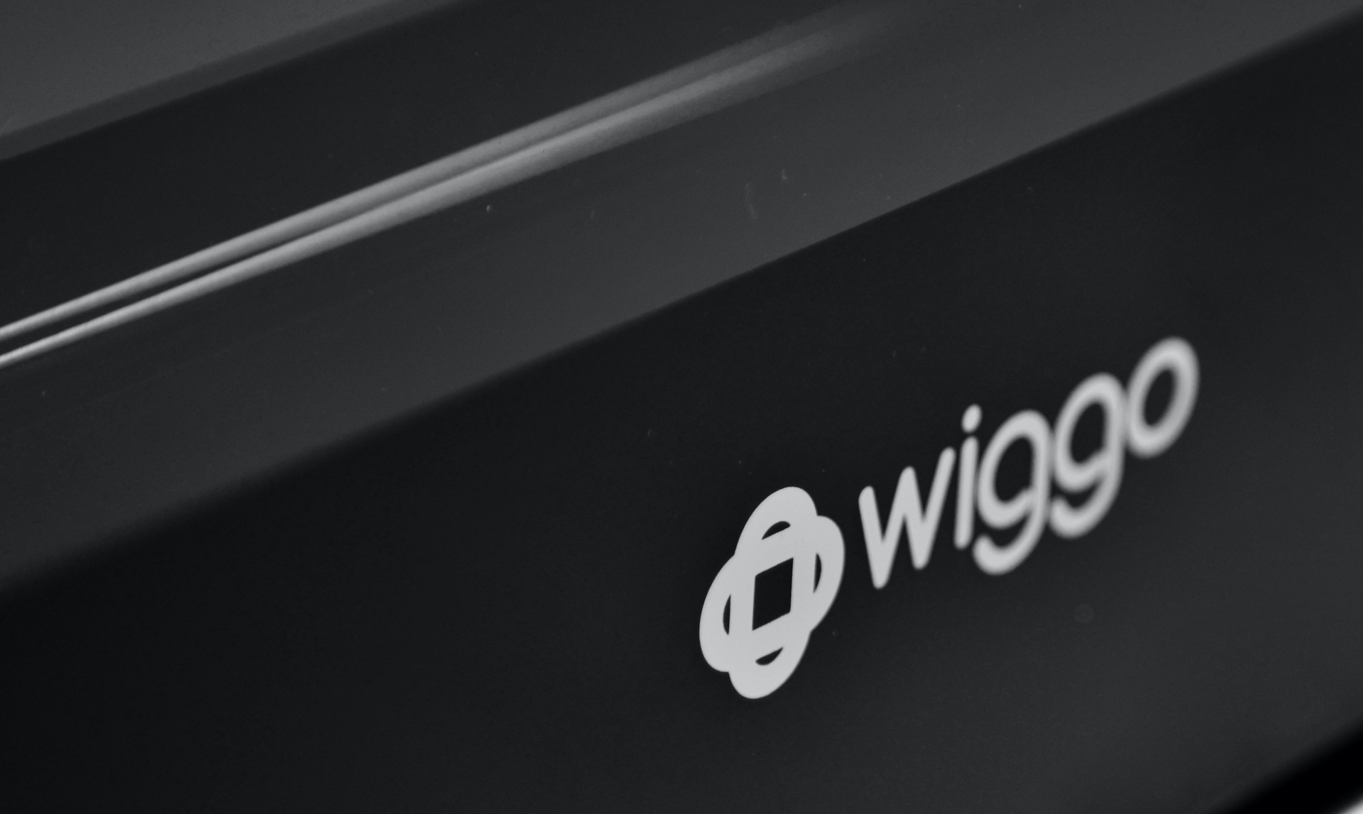 Gros plan d'une surface noire avec le logo blanc 'wiggo'. La surface a un éclat réfléchissant et deux lignes claires.