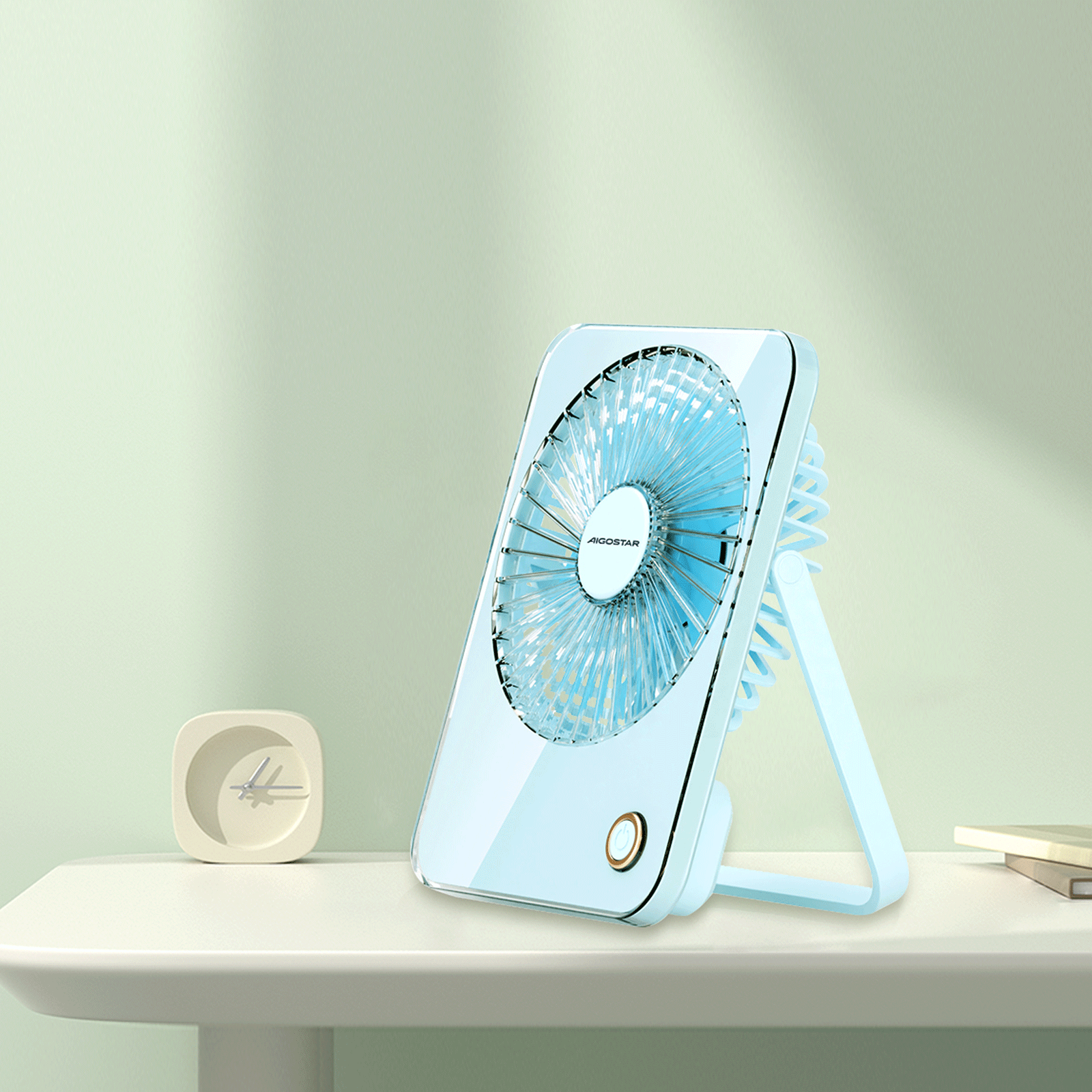 Un ventilador portátil azul con la marca AIGOSTAR está sobre una mesa blanca. También hay un reloj pequeño y libros.