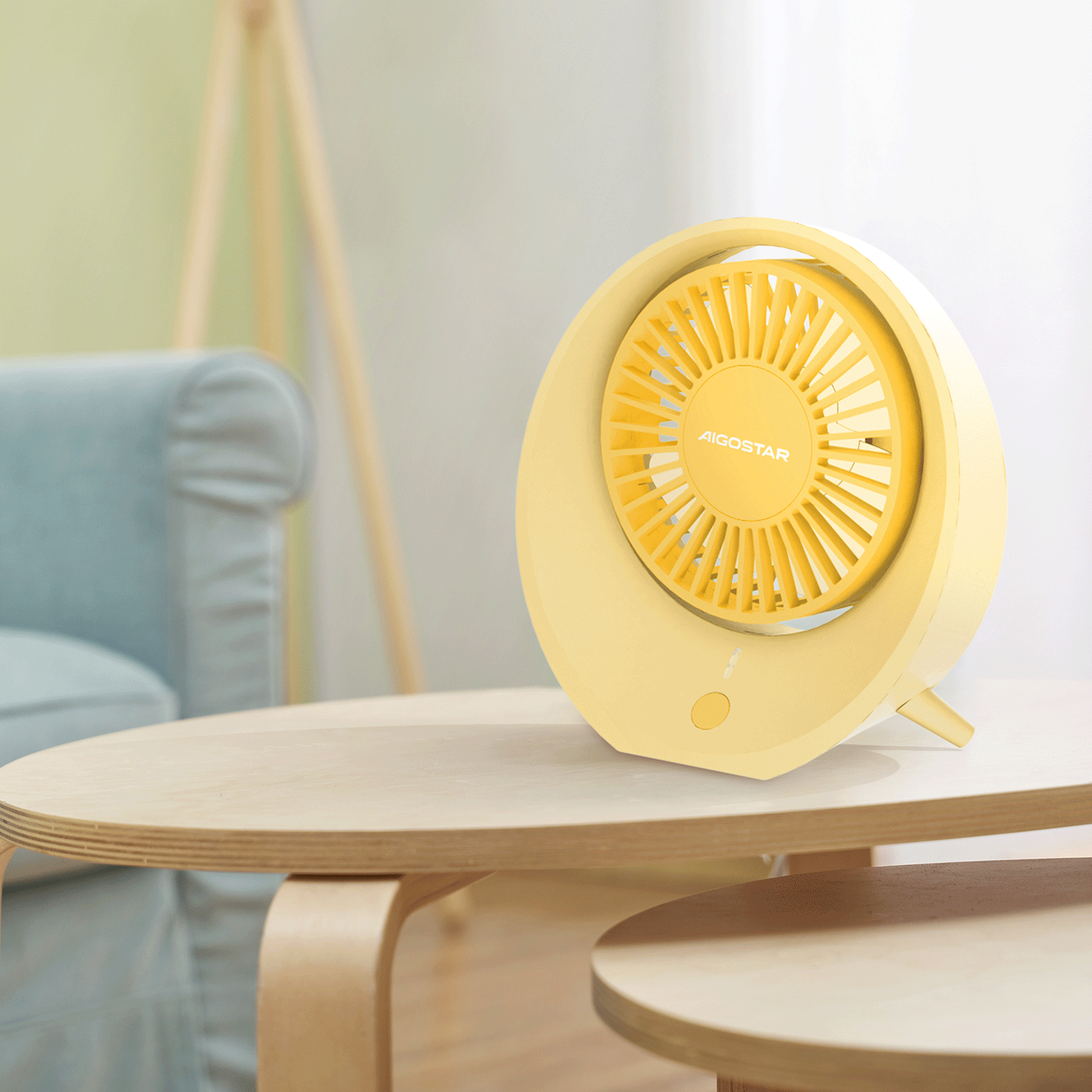 Ventilatore giallo Aigostar su un tavolo di legno. Divano blu chiaro e cornice di legno sullo sfondo sfocato.