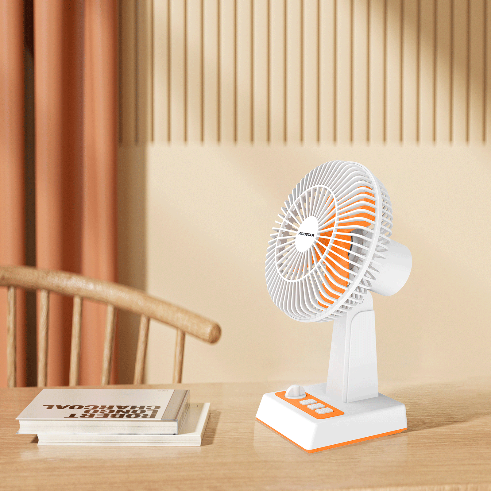 Un ventilador blanco y naranja está sobre una mesa de madera. También hay un libro y una silla.