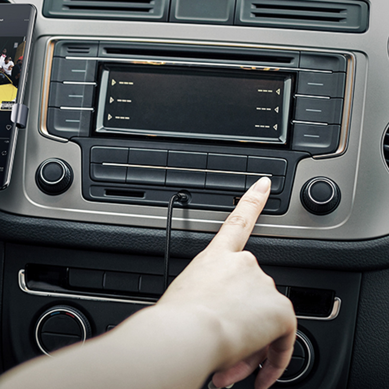 Dłoń naciska przycisk w radiu samochodowym. Telefon jest zamontowany po lewej stronie. Radio ma ekran i przyciski.