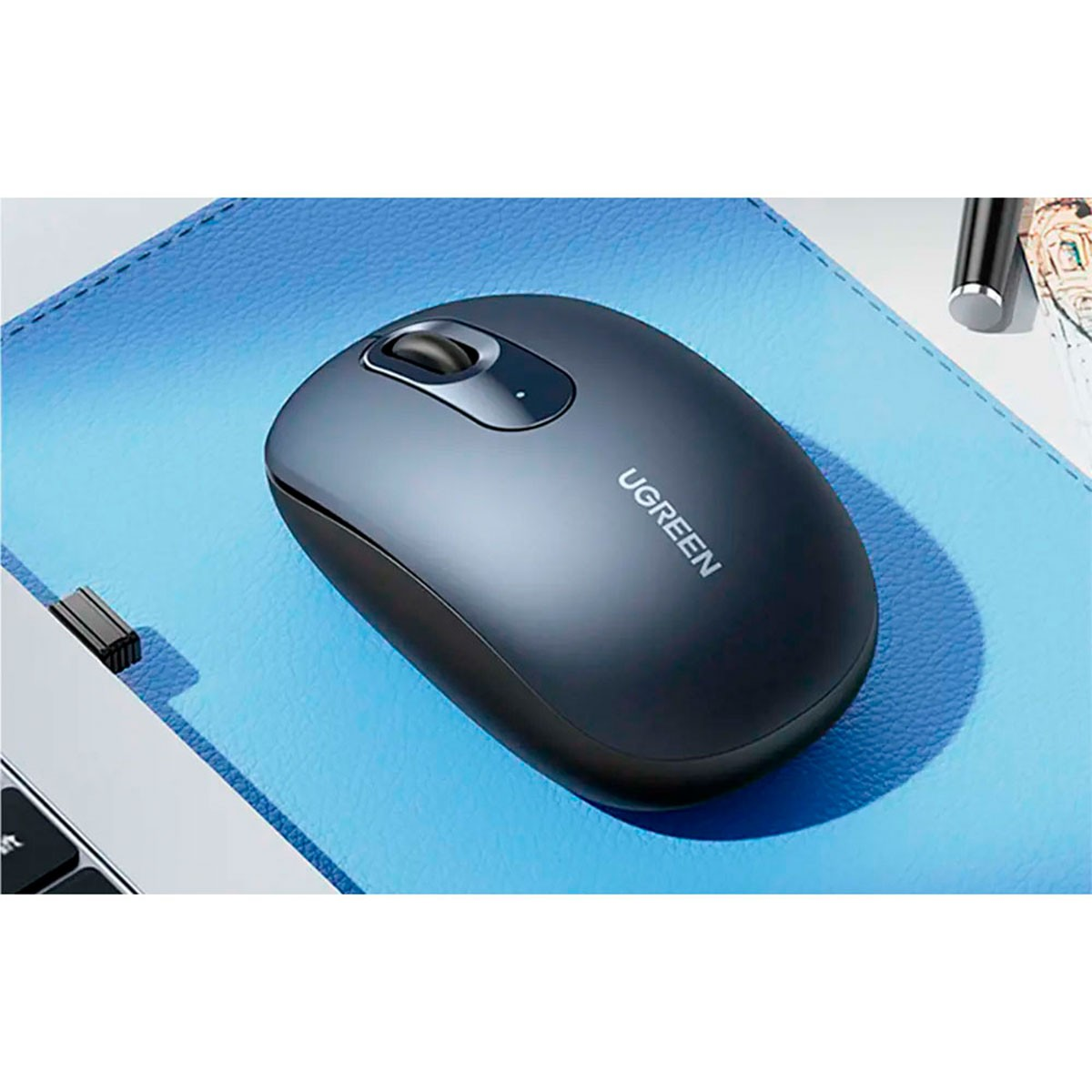 Ciemnoszara mysz UGREEN na niebieskiej podkładce pod mysz, z laptopem i długopisem w tle.