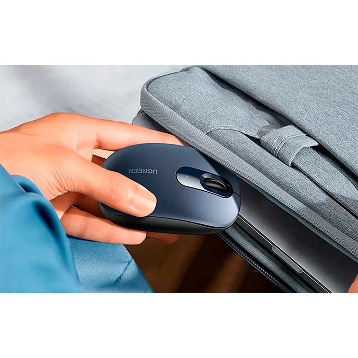 Ręka trzyma niebieską mysz UGREEN obok szarej torby na laptopa, sugerując przenośność i integrację z technologią.