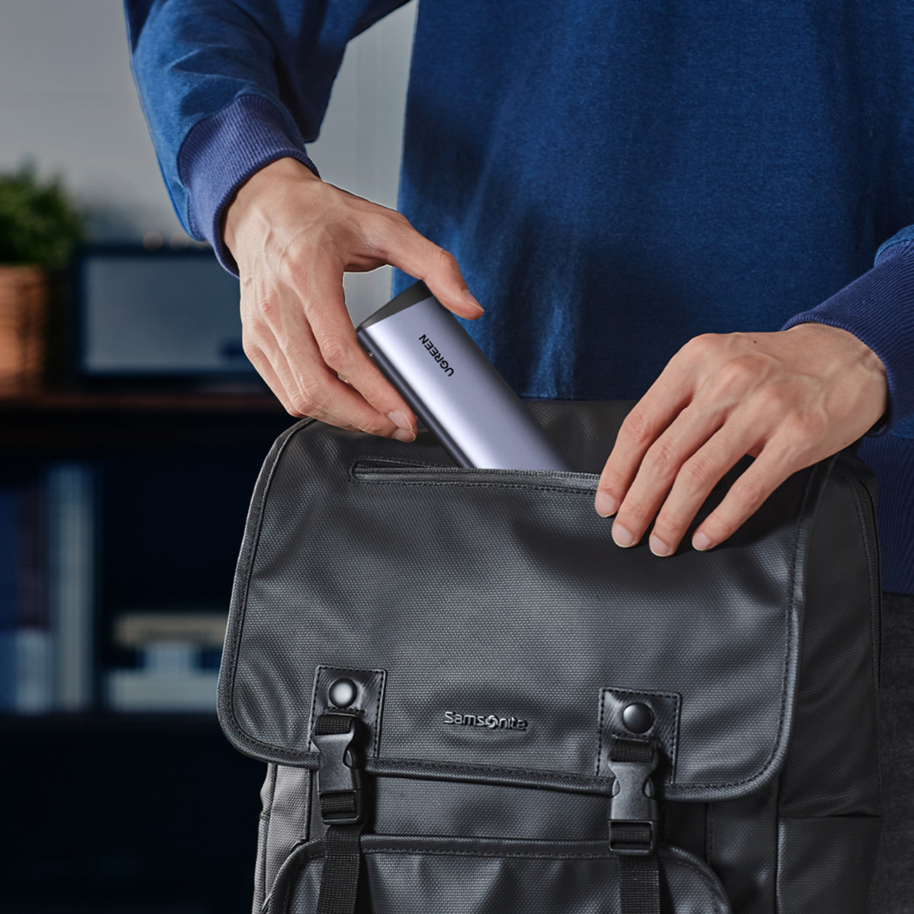 Osoba wkłada szare urządzenie UGREEN do czarnego plecaka Samsonite. Osoba ma na sobie niebieski sweter. Regał z książkami w tle.