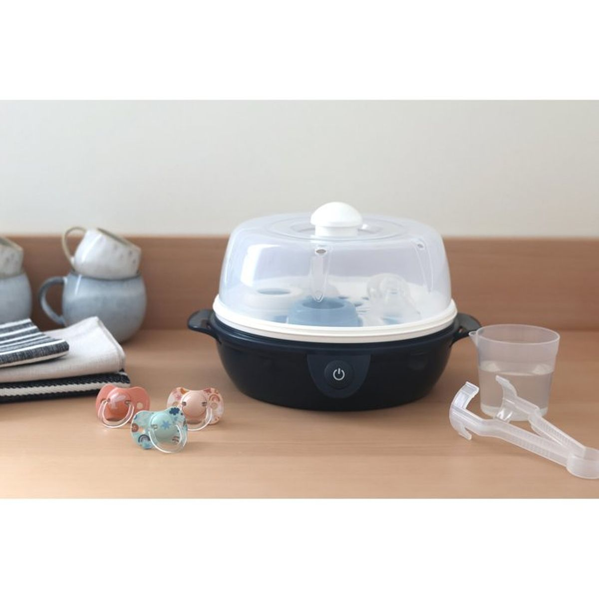 Ein schwarzer Babyflaschensterilisator mit Schnullern, Wasserbecher und Zange auf einer Holzoberfläche. Tassen und Handtücher sind im Hintergrund.