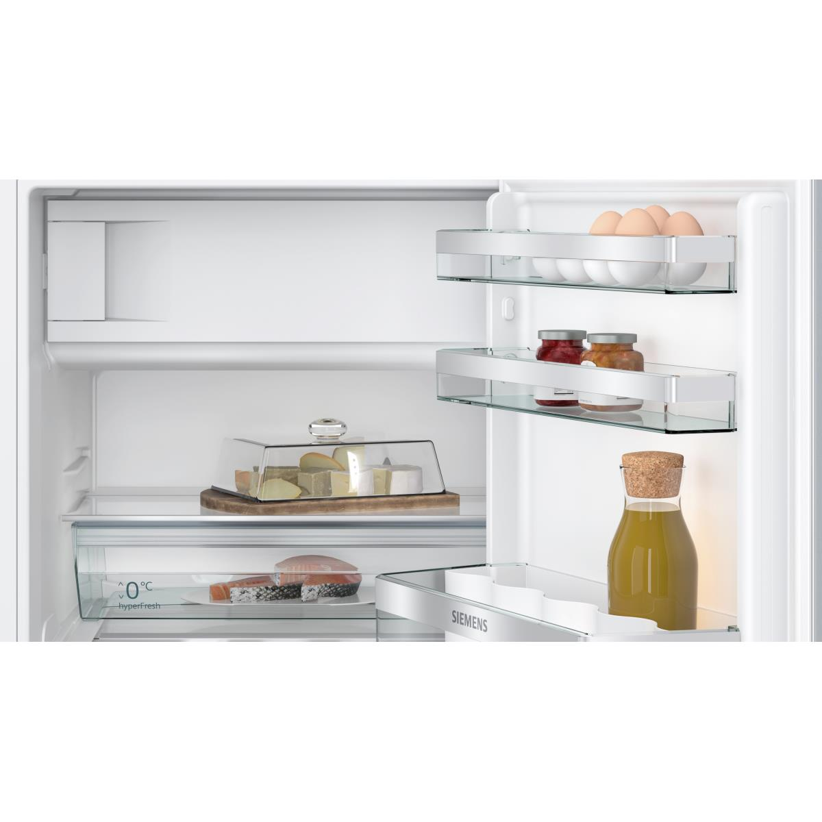 Im Kühlschrank: Käse, Lachs, Gläser, Eier und eine Ölflasche sind zu sehen. Die Regale sind aus Glas, der Kühlschrank ist weiß.