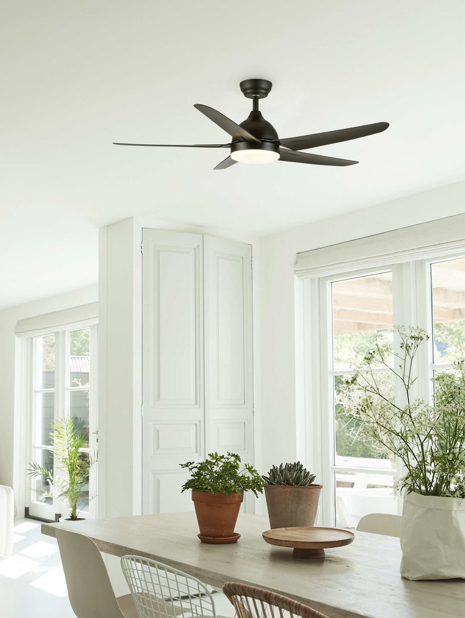 Sala da pranzo luminosa con ventilatore a soffitto. Tavolo con piante e vassoio in legno. Finestre e pareti bianche.
