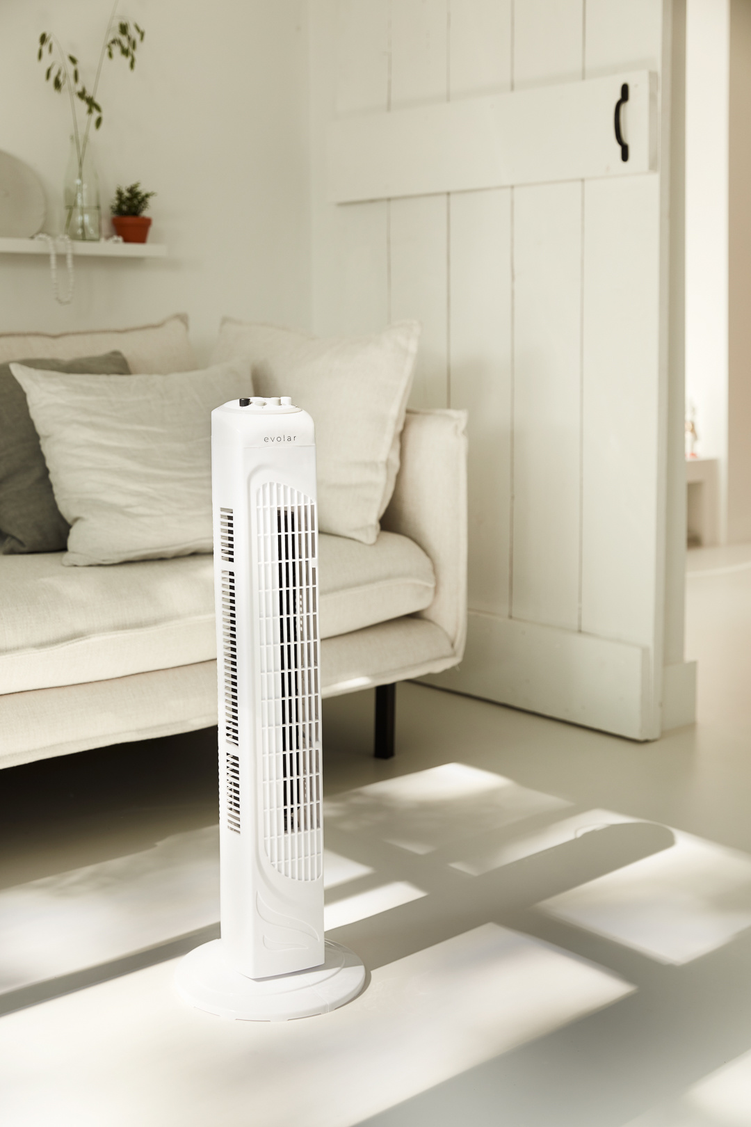 A white tower fan stands on a white floor, near a cream-colored sofa. Sunlight streams in, casting shadows.