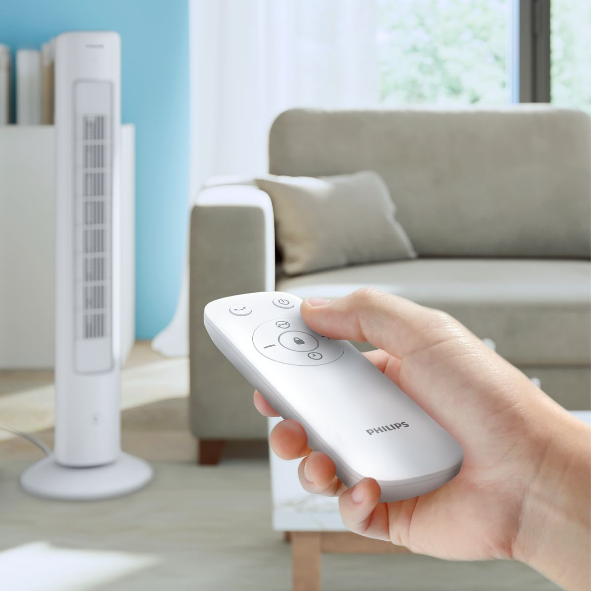 Ręka trzyma biały pilot Philips, kierując go w stronę białego wentylatora. W tle sofa i okno.