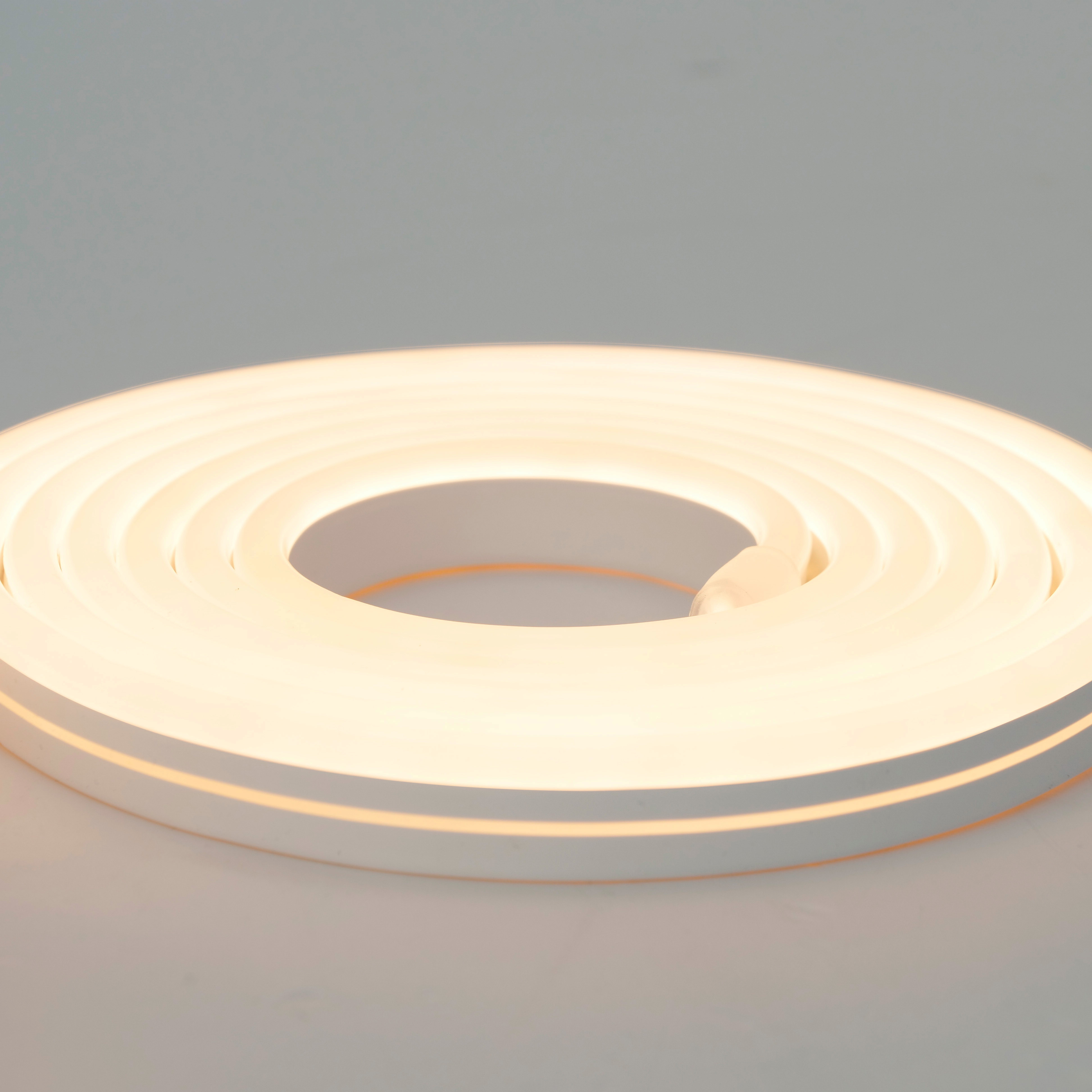 Eine aufgerollte, beleuchtete Neonlichtleiste, die ein warmes Leuchten ausstrahlt. Der Hintergrund ist hellgrau.